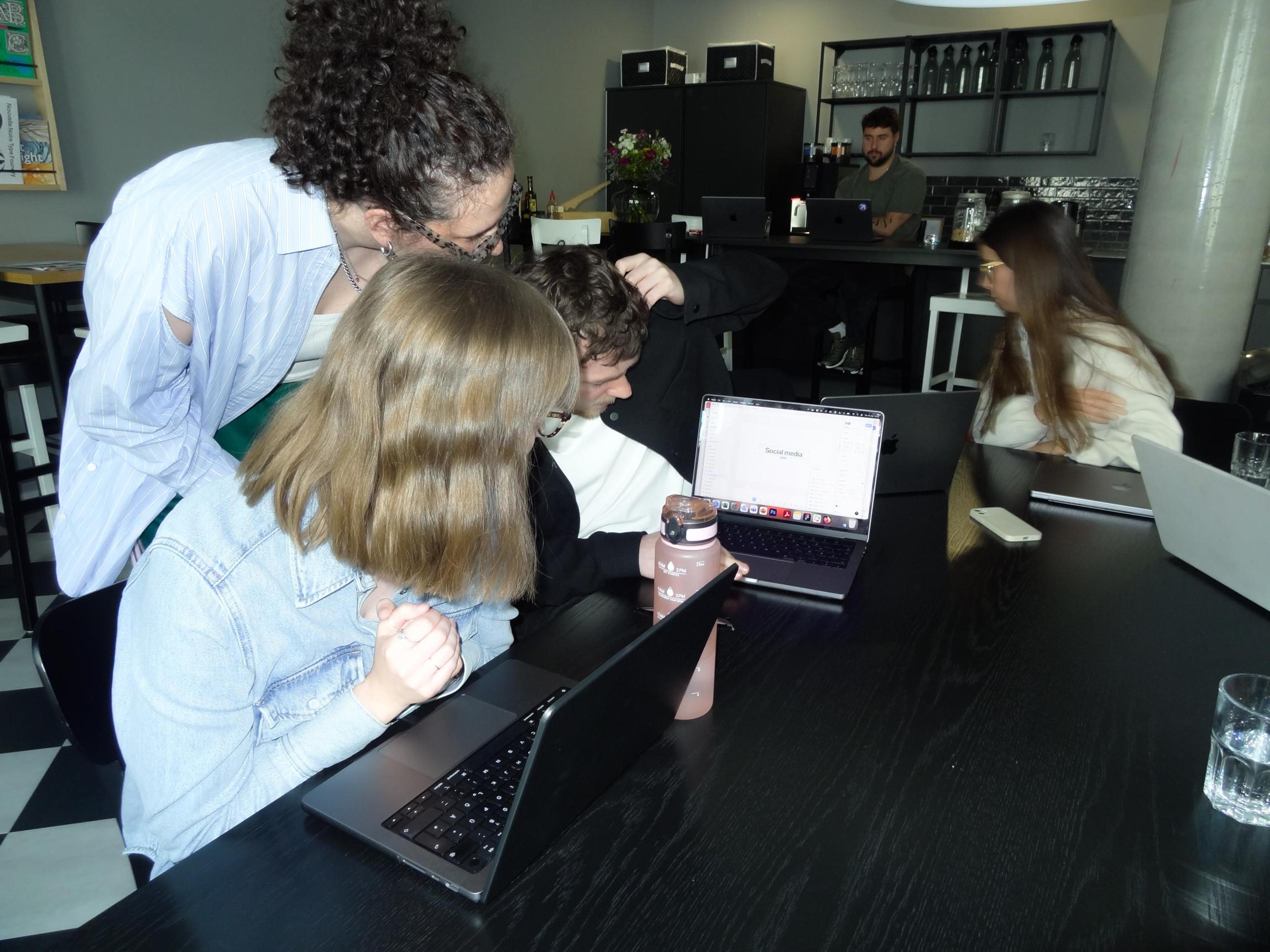 A group of four people gathered around a black table, focusing on laptop screens. The laptops display information, with one showing "Social media" text. Visible expressions suggest concentration and collaboration. Additional individuals are seated further back in the space, also using laptops.
