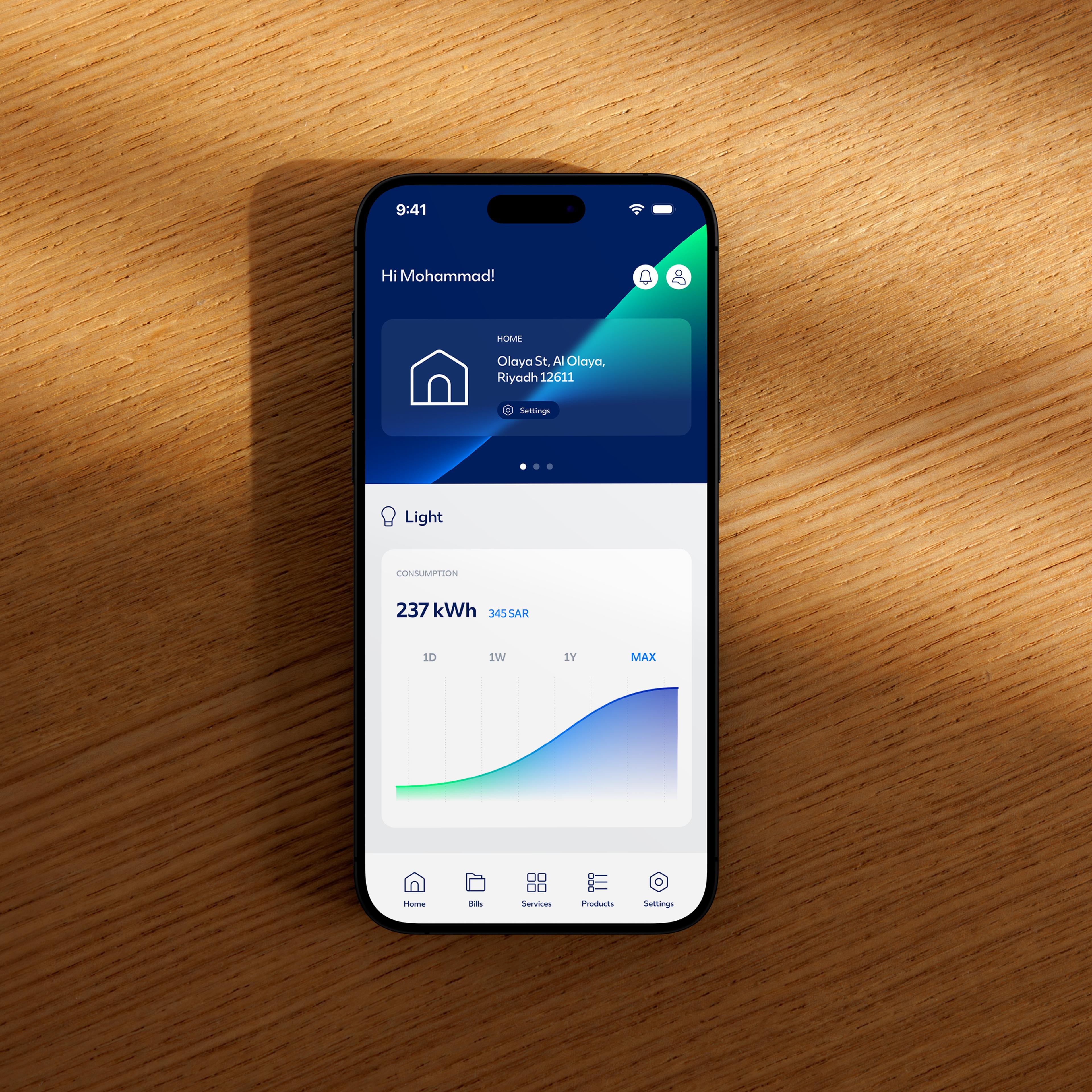 Modern smartphone displaying a smart home energy management app on a wooden table, featuring a blue dashboard interface with the greeting “Hi Mohammad!” and home address “Olaya St, Al Olaya, Riyadh 12611.” Screen shows electricity consumption statistics of 237 kWh costing 345 SAR, with an upward trend graph labeled 1D, 1W, 1Y, MAX. Bottom navigation icons include Home, Bills, Services, Products, and Settings, highlighting a digital utility monitoring app, electricity usage analytics, and smart home dashboard UI design.