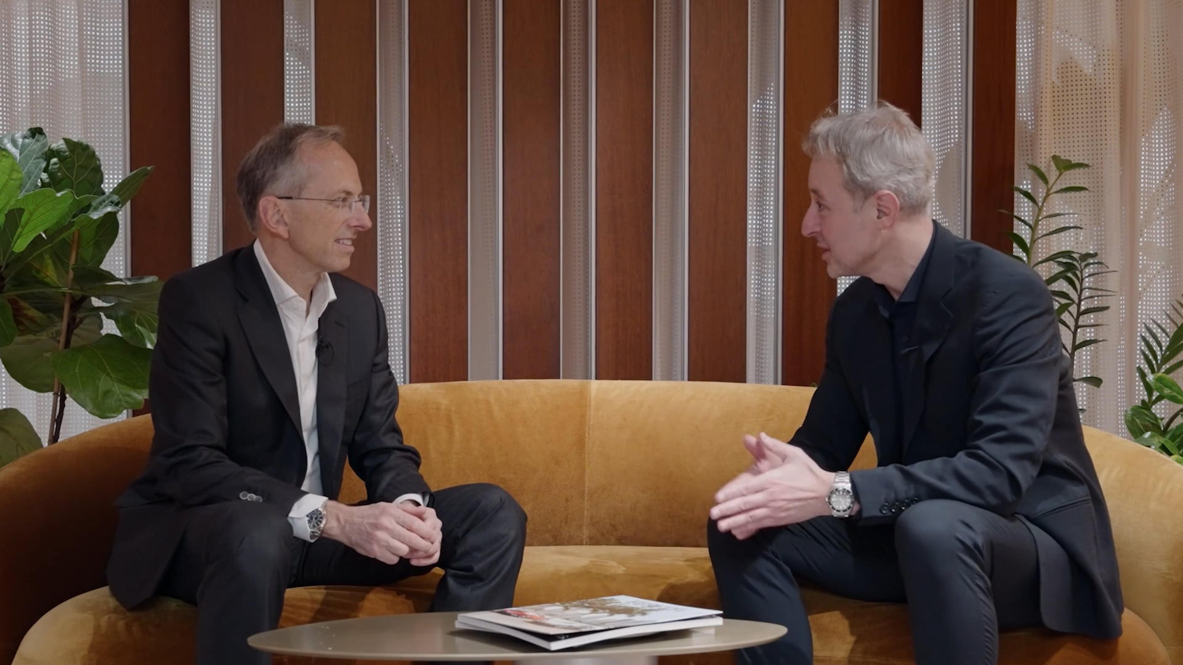 Benedetto Vigna and Manfredi Ricca sit on a yellow sofa having a conversation. A round coffee table with magazines is in front of them. The setting is warm with plants and wood paneling.