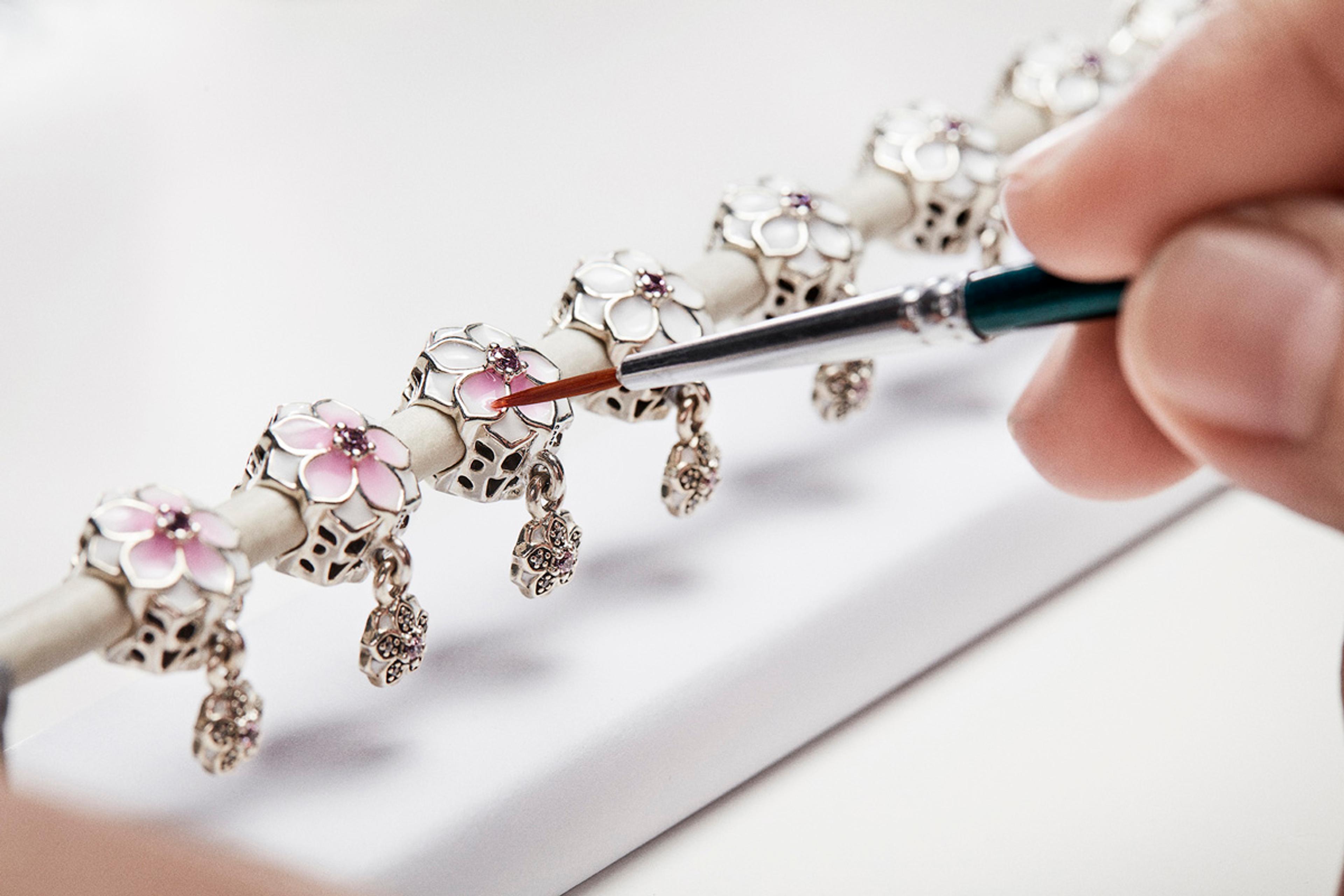 A craftsperson applies enamel paint with a fine brush to a silver Pandora charm bracelet displayed on a white cylindrical stand. The bracelet features multiple floral charms with pink and white enamel petals, purple gemstone centres, and dangling heart-shaped pendants.