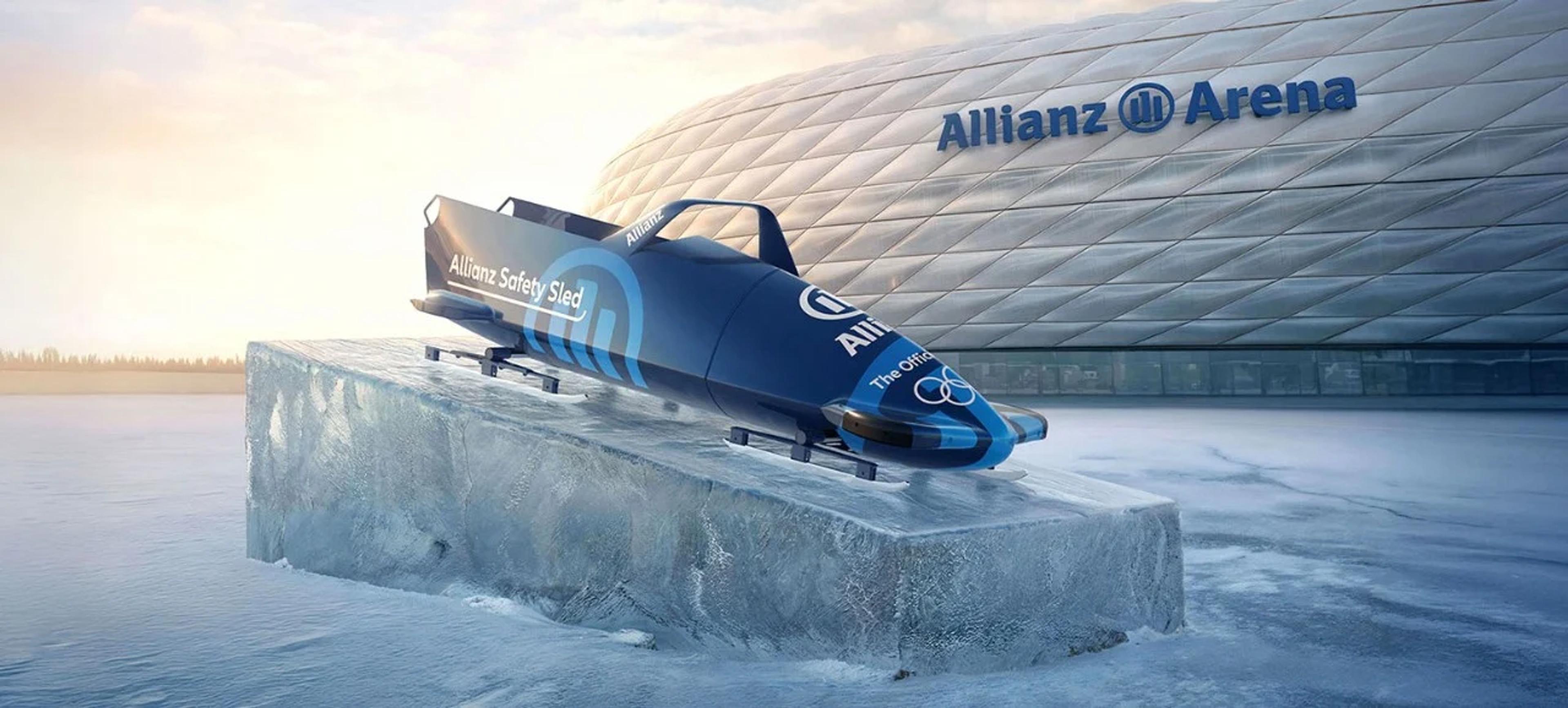 A CGI-rendered navy blue bobsled branded "Allianz Safety Sled" sits on a large block of ice in front of the Allianz Arena in Munich. The sled features the Allianz logo and Olympic rings. The scene has a cold, wintry atmosphere with a pale golden sky in the background.