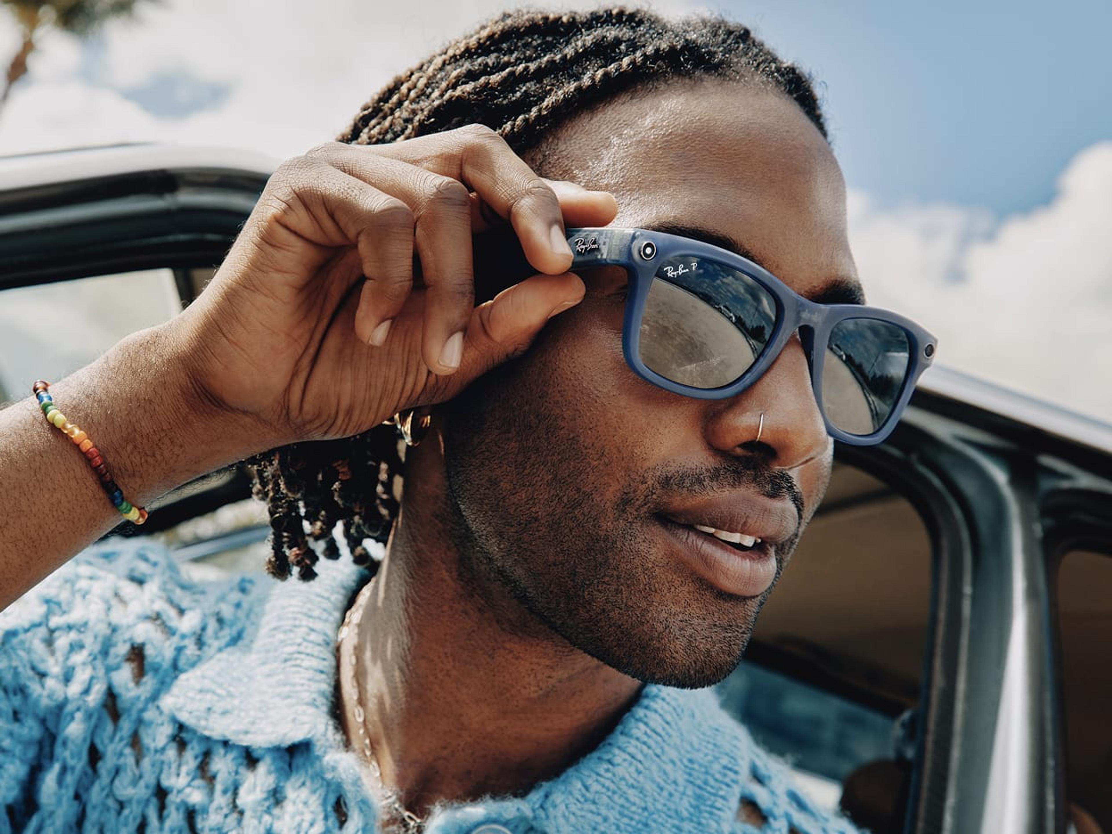 Close-up of a man adjusting a pair of blue Ray-Ban Meta smart glasses, with a small camera visible on the temple. He wears a blue chunky-knit top, a rainbow beaded bracelet, and a nose ring, and is seated in an open-top car against a bright sky.