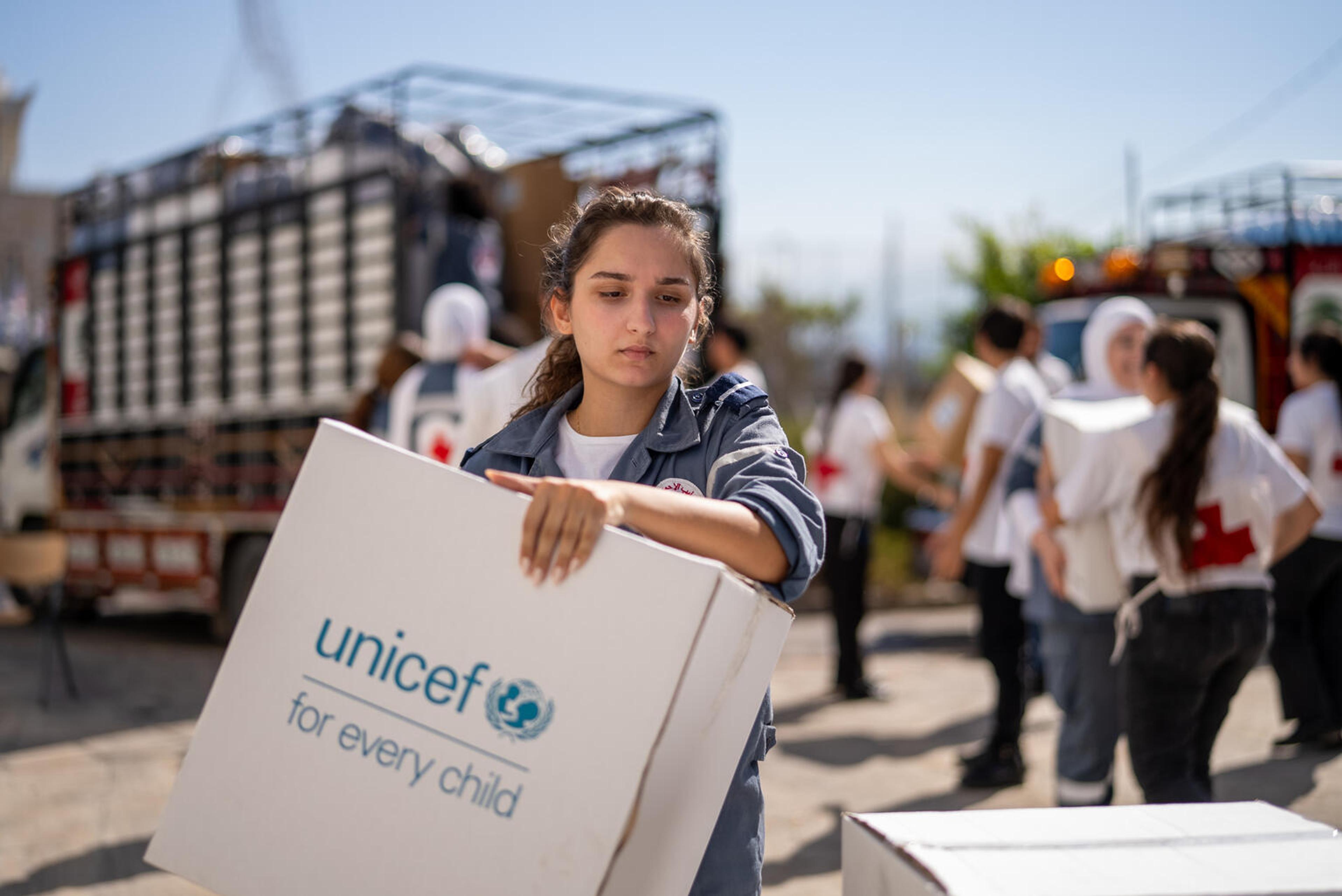 A young woman in a grey jacket handles a white UNICEF supply box labelled "for every child" during an outdoor aid distribution. In the background, a group of volunteers wearing Red Cross vests unload cargo from large trucks under a clear sky.