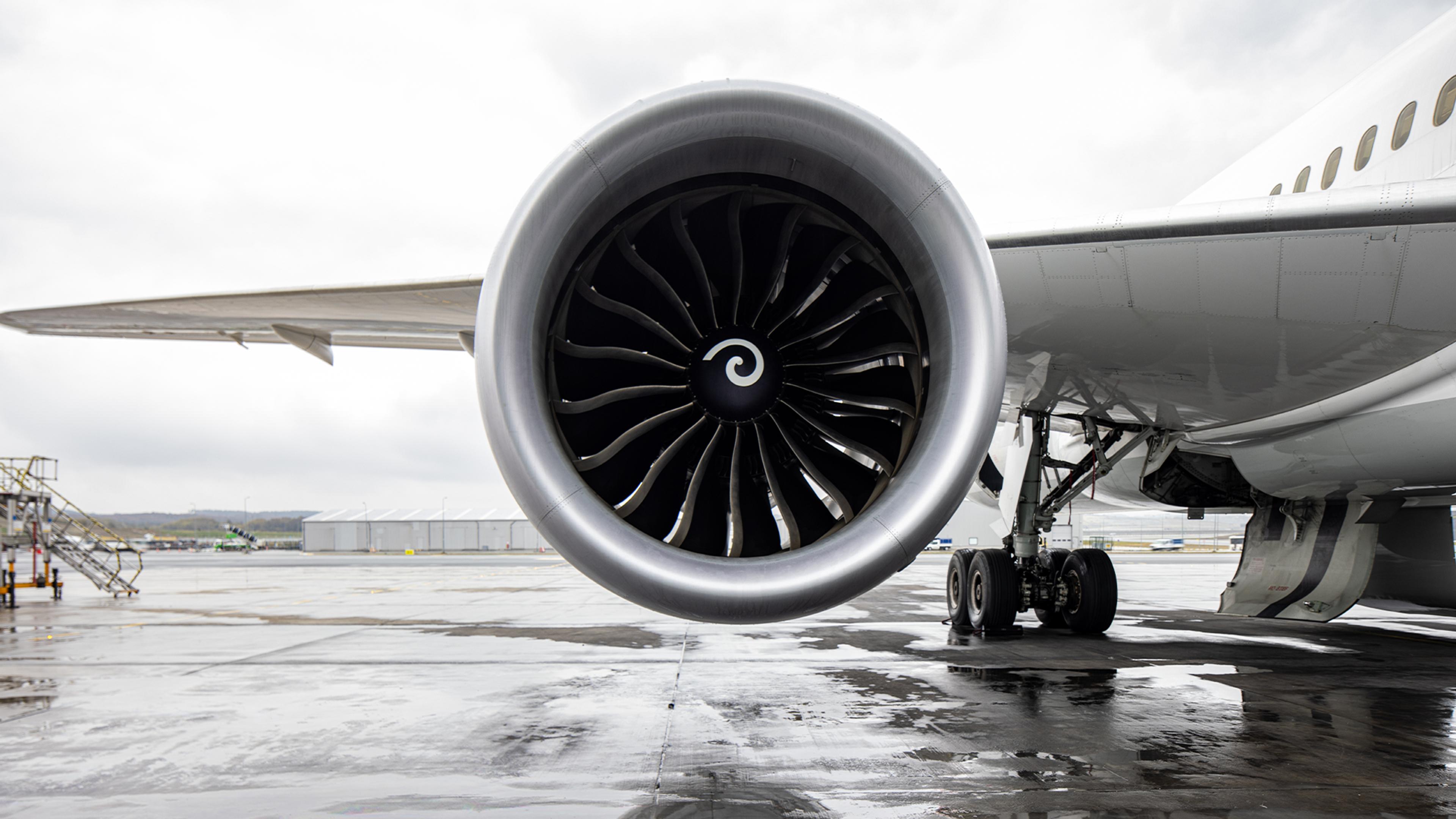Close-up ground-level view of a GEnx jet engine mounted under the wing of a white commercial aircraft, parked on a wet tarmac on an overcast day. The engine's large fan blades and spiral spinner are sharply in focus, with the aircraft's landing gear and airport facilities visible in the background.