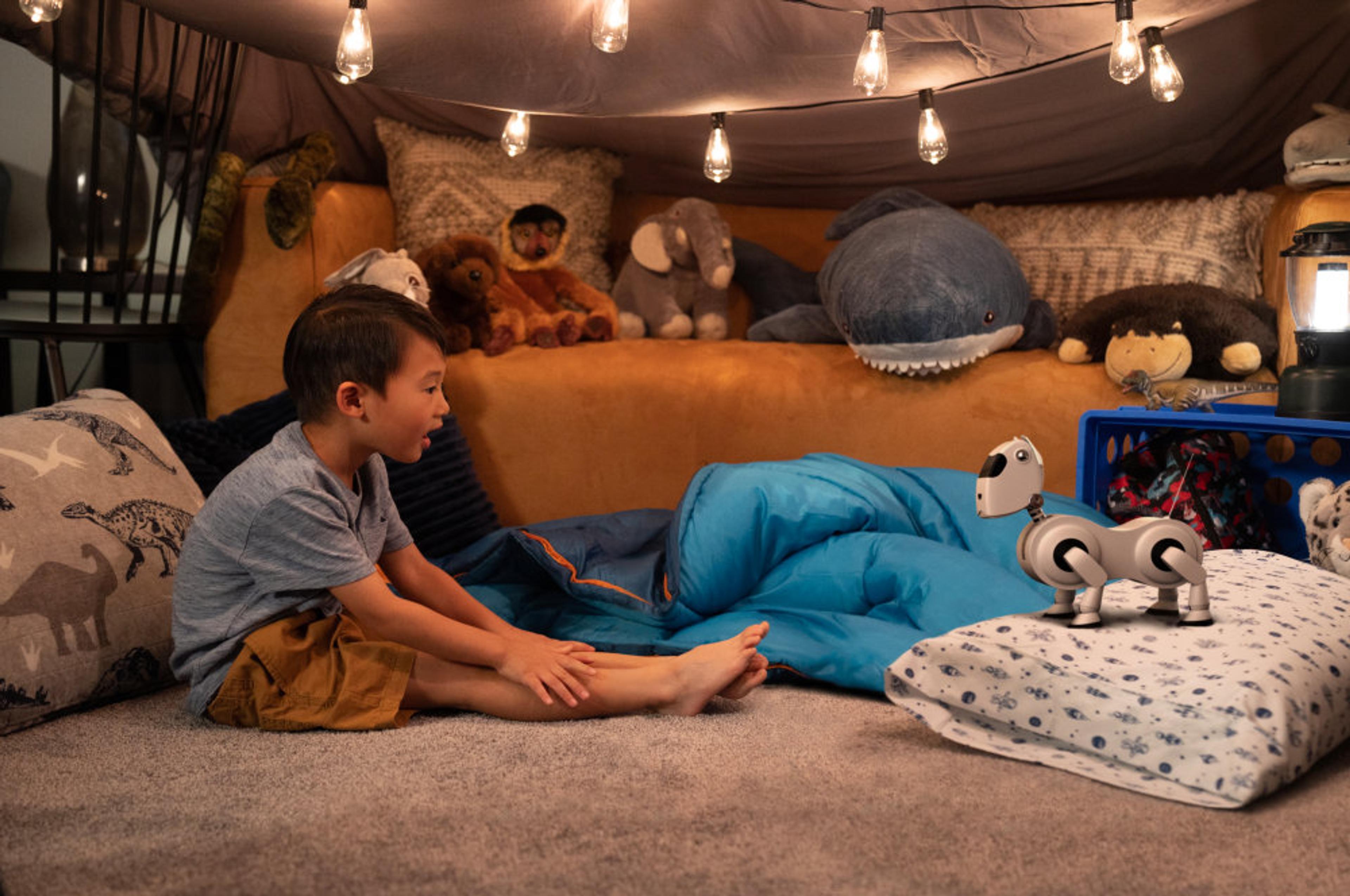 A young boy sits cross-legged on the floor of a cosy indoor blanket fort, looking with curiosity at a small white Sony Aibo robot dog standing nearby. The fort is lit by warm string lights and surrounded by stuffed animals, a blue sleeping bag, and dinosaur-print pillows.