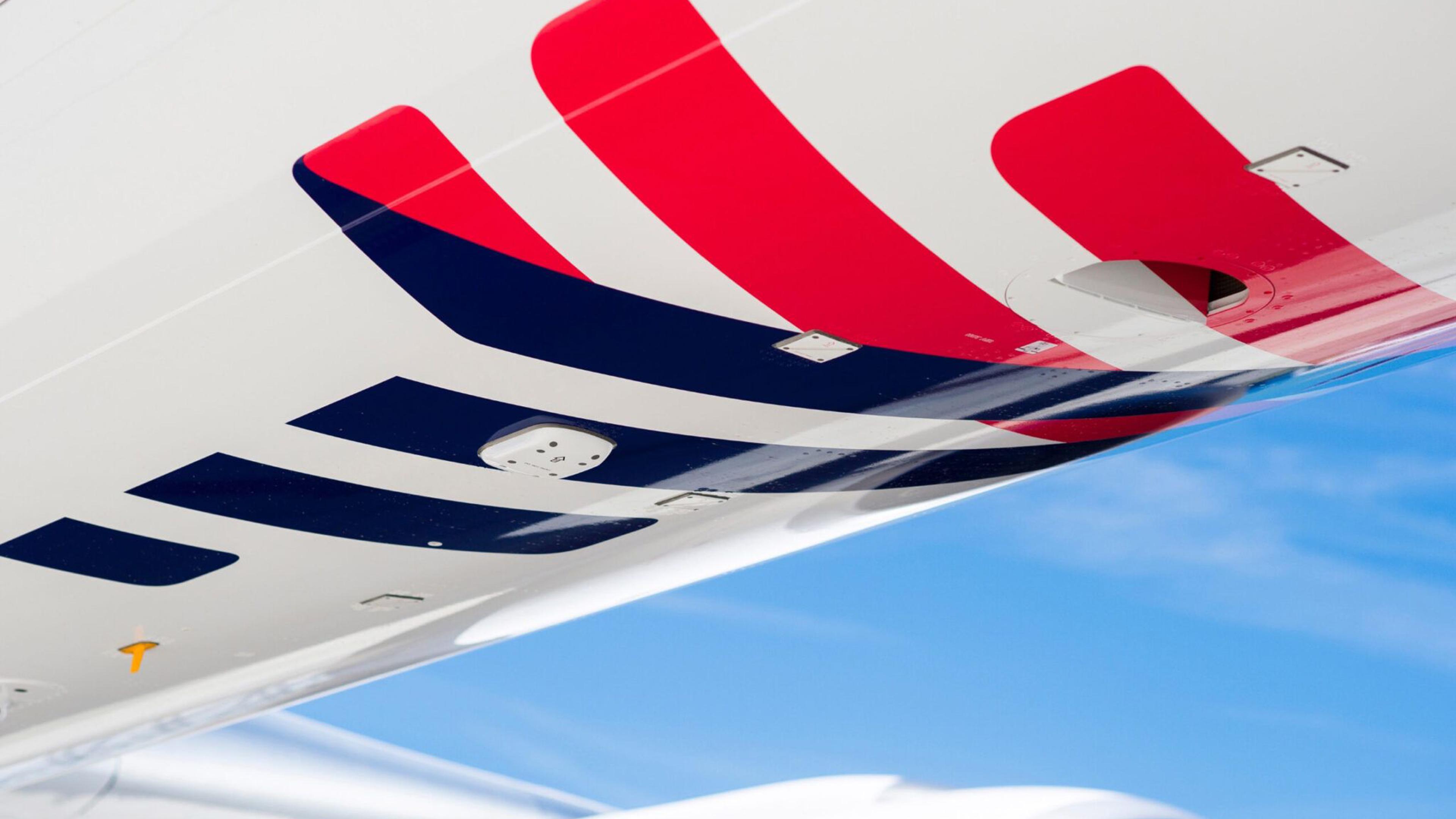 Close-up view of the upper portion of an aircraft wing featuring a white surface with red and blue stylized stripes. The wing houses a sensor, and a portion of the blue sky and the engine cowling are visible in the background.