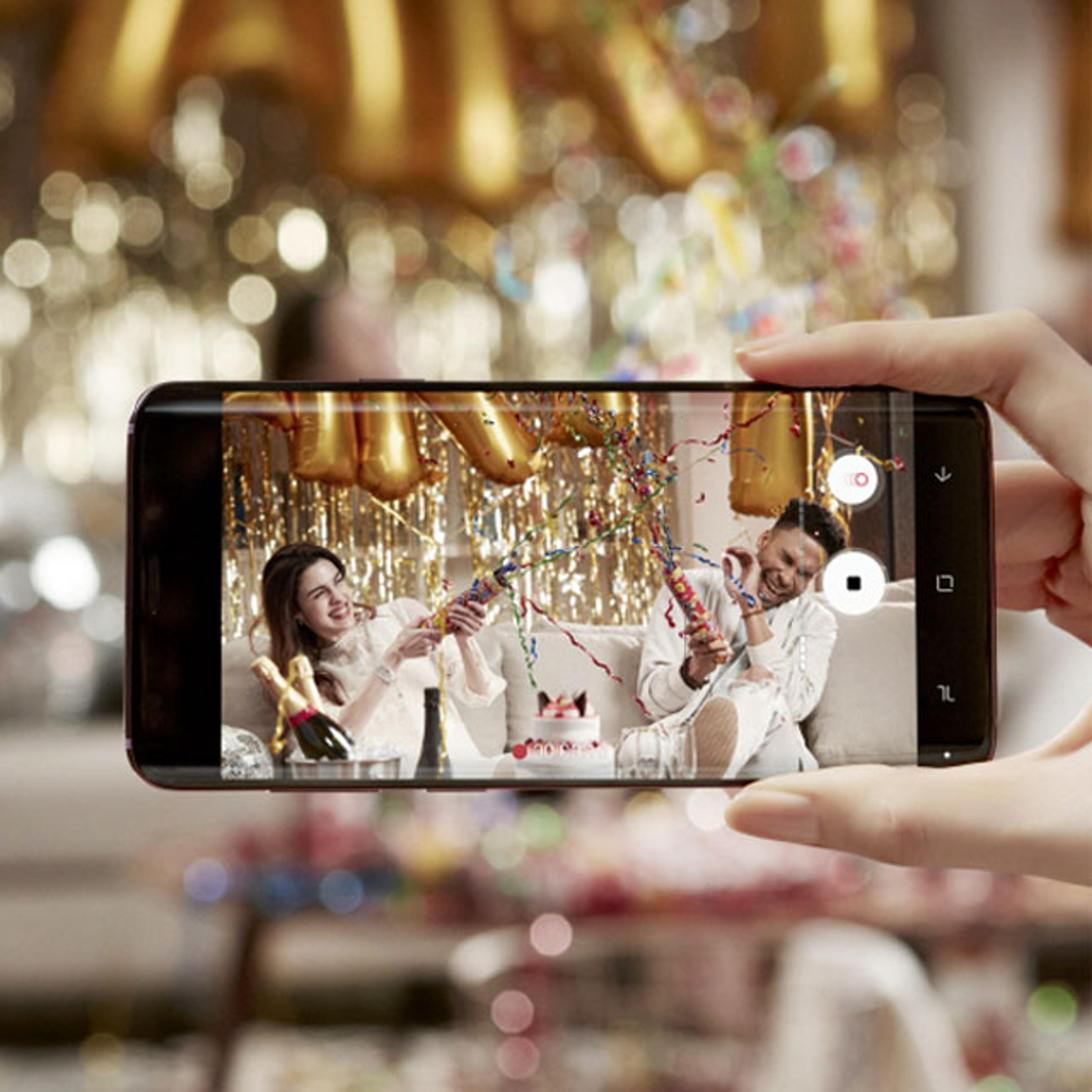 A person holds a Samsung phone taking a photo of two people celebrating with party poppers, surrounded by gold decorations and a cake.