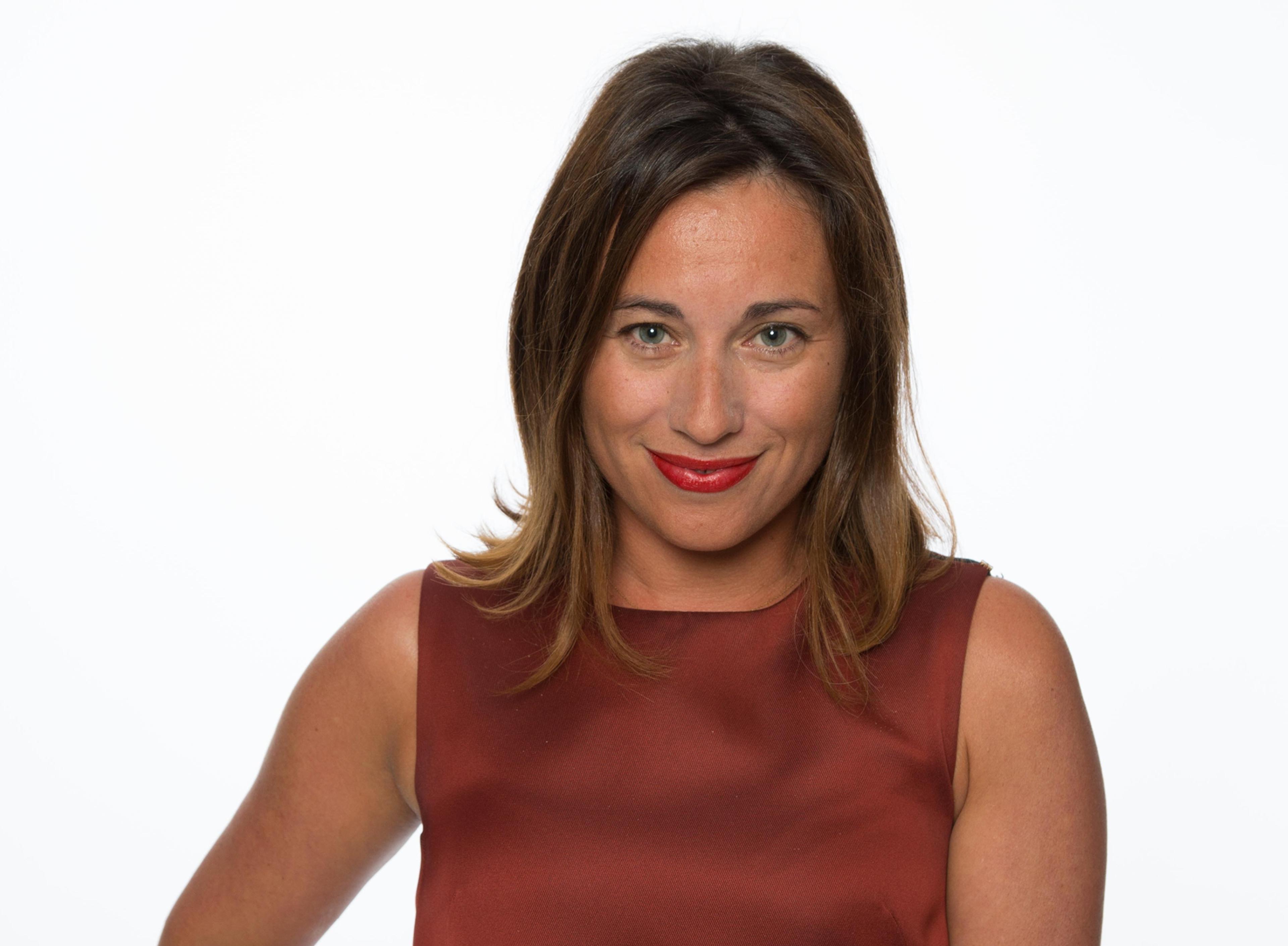 A woman poses. She has mid-length brown hair and red lipstick. She wears a deep red top.
