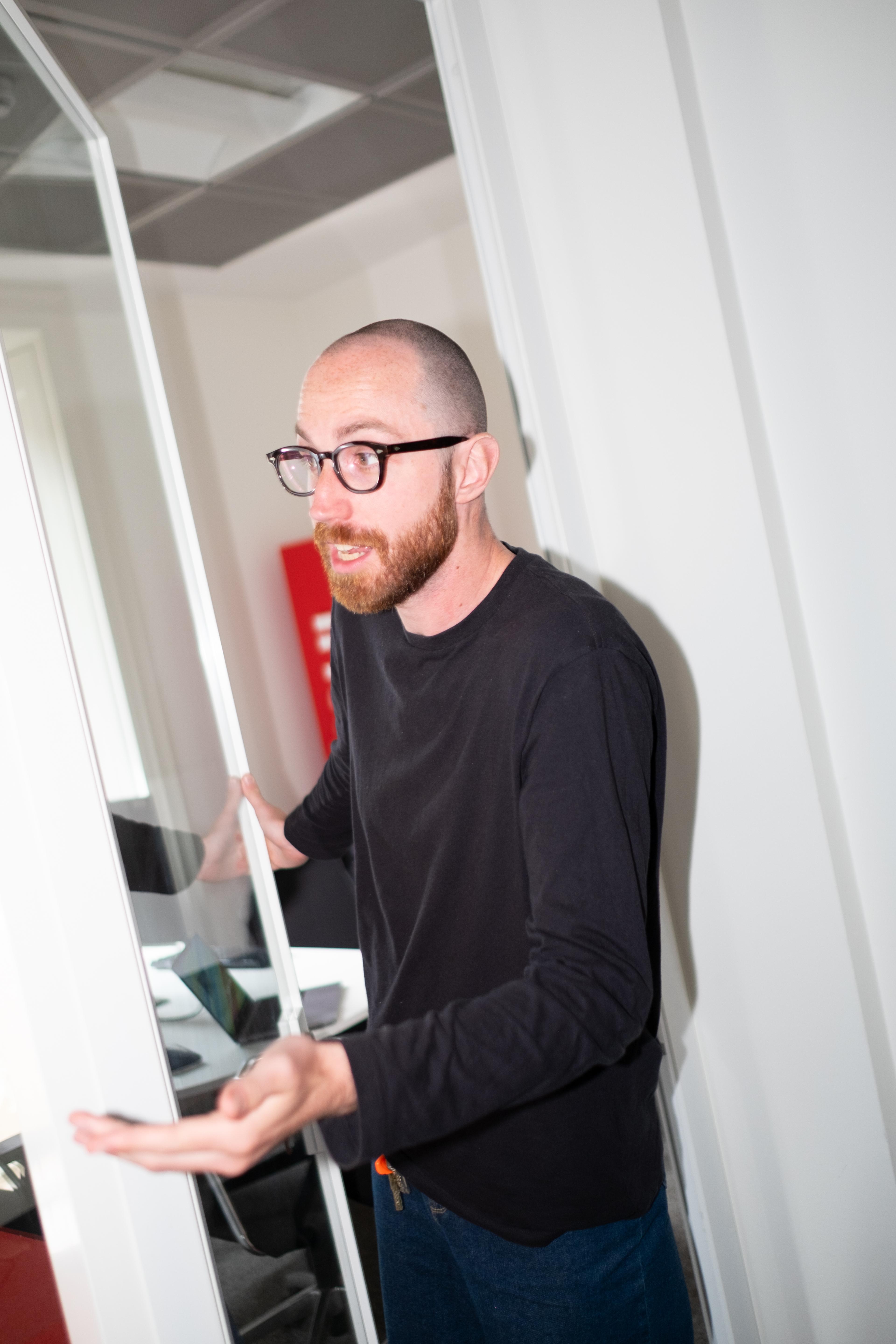 A man in glasses and a black shirt gestures while presenting in a modern office, with a large mirror or glass panel beside him.